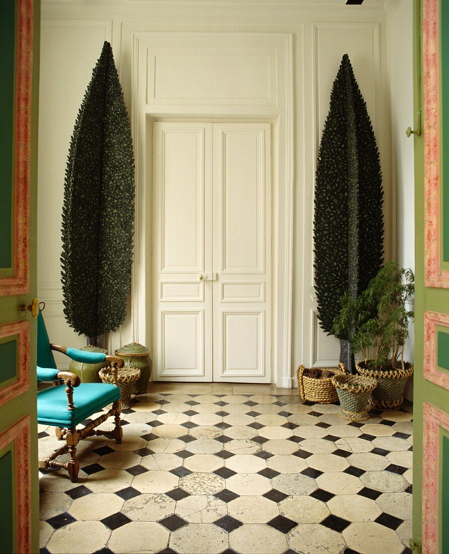 Stilisierte Bäume zu beiden Seiten einer Flügeltür in Vorraum mit schwarzweiss gefliestem Boden und antikem, türkisfarbenen Armlehnstuhl