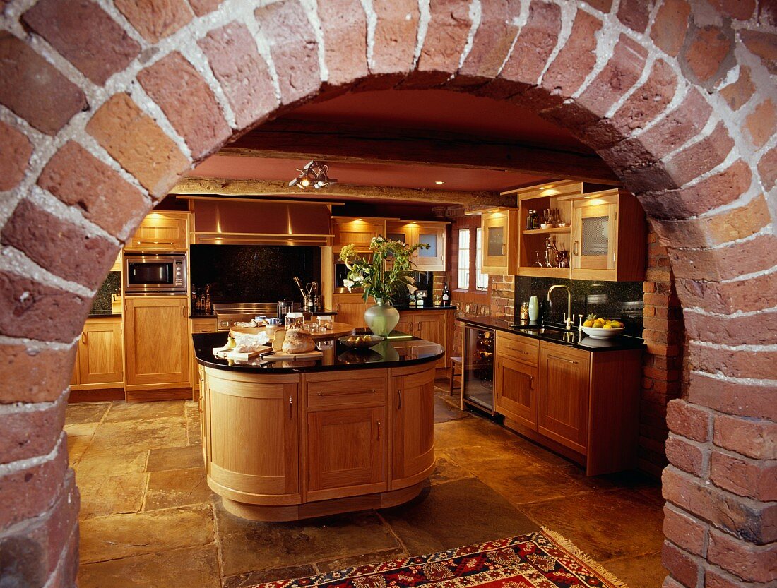 Blick durch rustikales Backsteingewölbe auf moderne Küche aus Holz mit ovaler Kochinsel und Steinboden