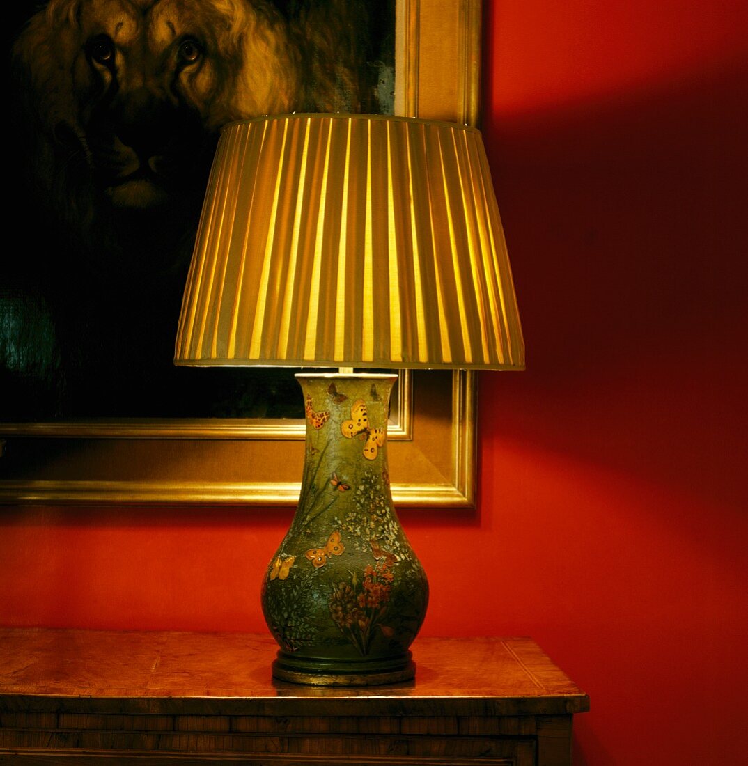 Brennende, antike Tischlampe mit bemaltem Porzellanfuss vor Ölgemälde auf roter Wand