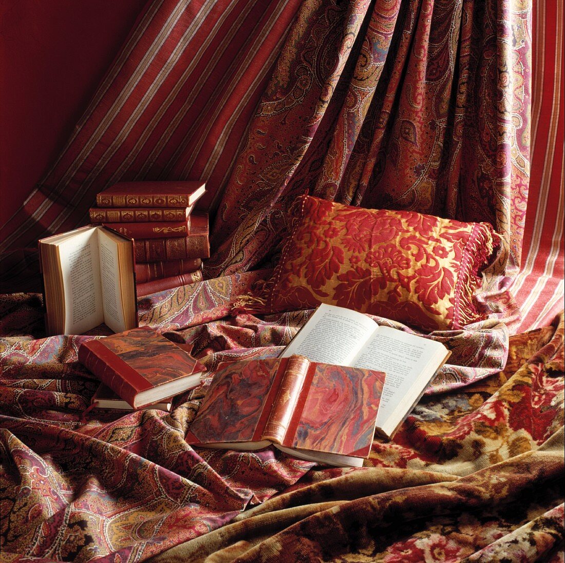 Alte Bücher mit marmoriertem Einband dekorativ drapiert auf rot/rosé und gold gemusterten Kissen und Vorhangstoffen