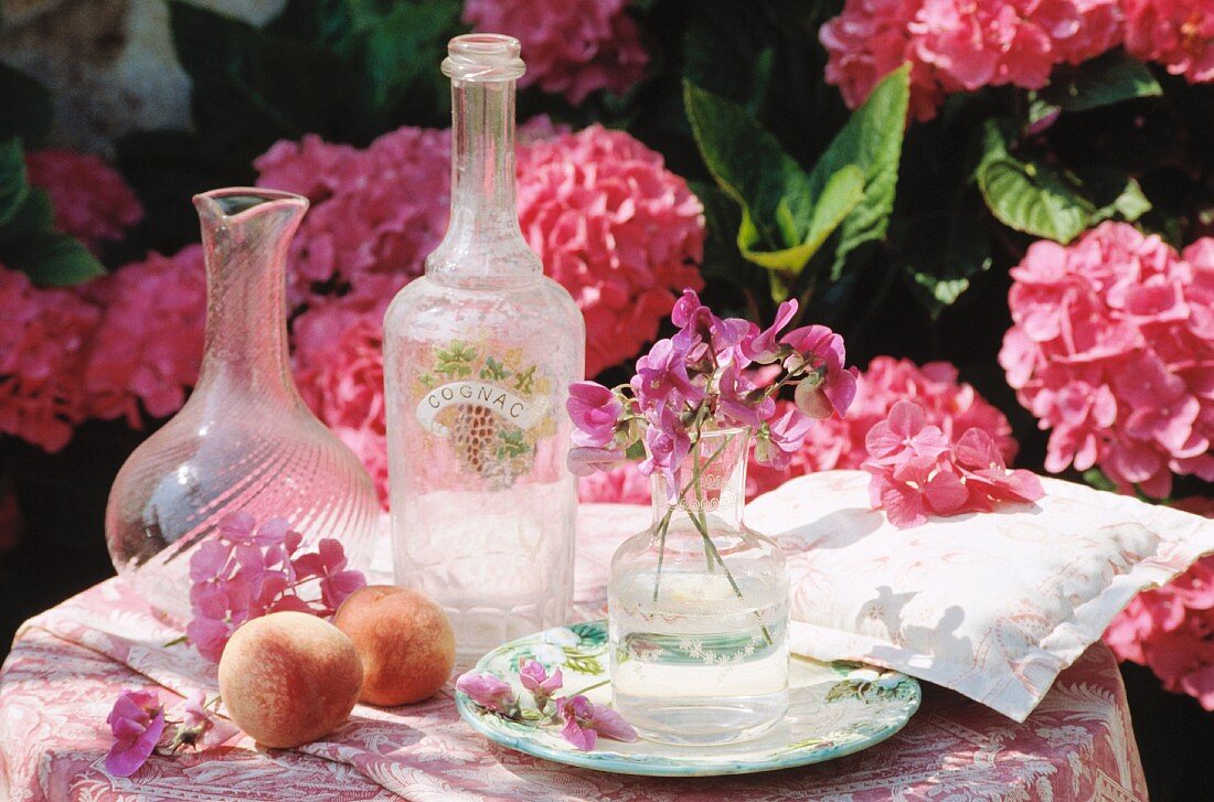 Glasflaschen auf Tisch mit rosé gemusterter Decke und Kissen vor rosa blühendem Hortensienbusch