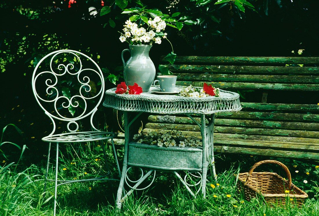 Stillleben in Wiese mit altem Metallstuhl und Blumenkrug auf Korbtisch