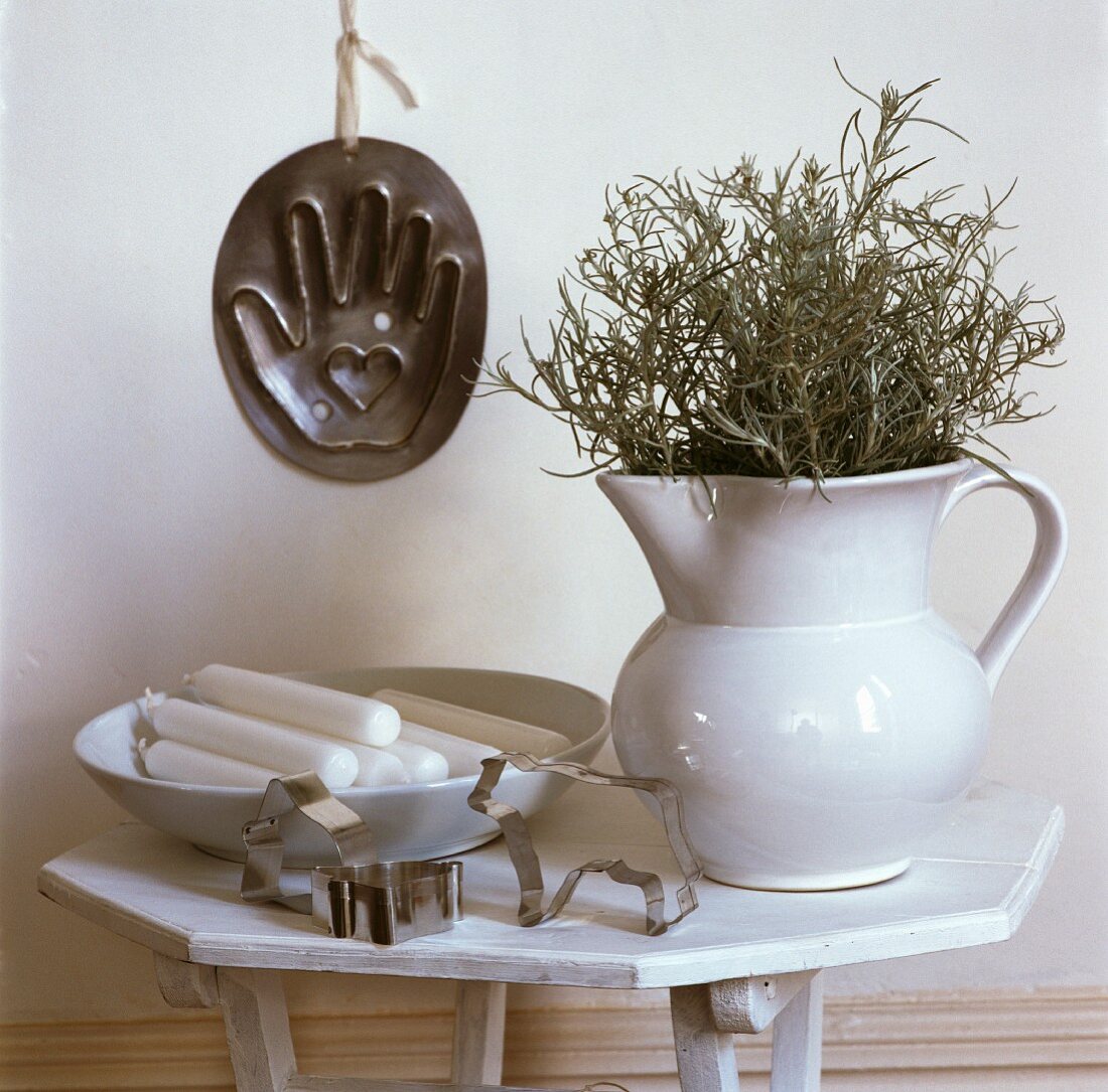 Hand-aufs-Herz-Backform an Wand hinter weißem Holztischchen mit Krug, Teller mit Kerzen und Ausstechformen