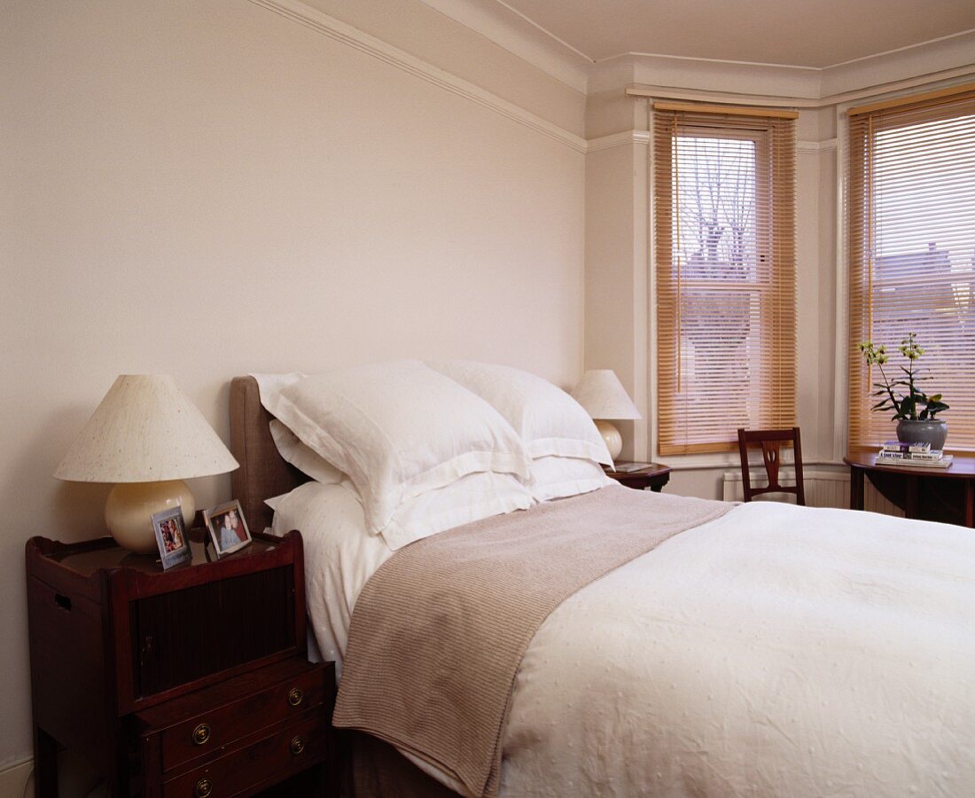 Erkerfenster mit Holzjalousien und antike Nachtkästchen neben weiss bezogenem Doppelbett in traditionellem Schlafzimmer