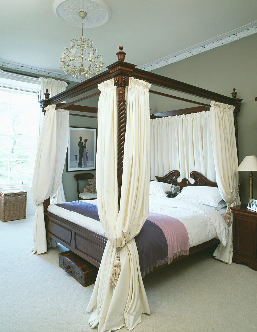 Cremefarbene Vorhänge an antikem Himmelbett aus dunklem Holz in traditionellem Schlafzimmer mit Kronleuchter