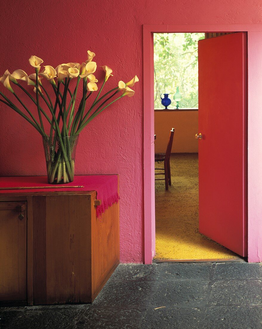 Vase mit Callas auf hölzernem Schrank in einem mexikanischen Saal mit leuchtend rosa Wänden und grauem Steinboden