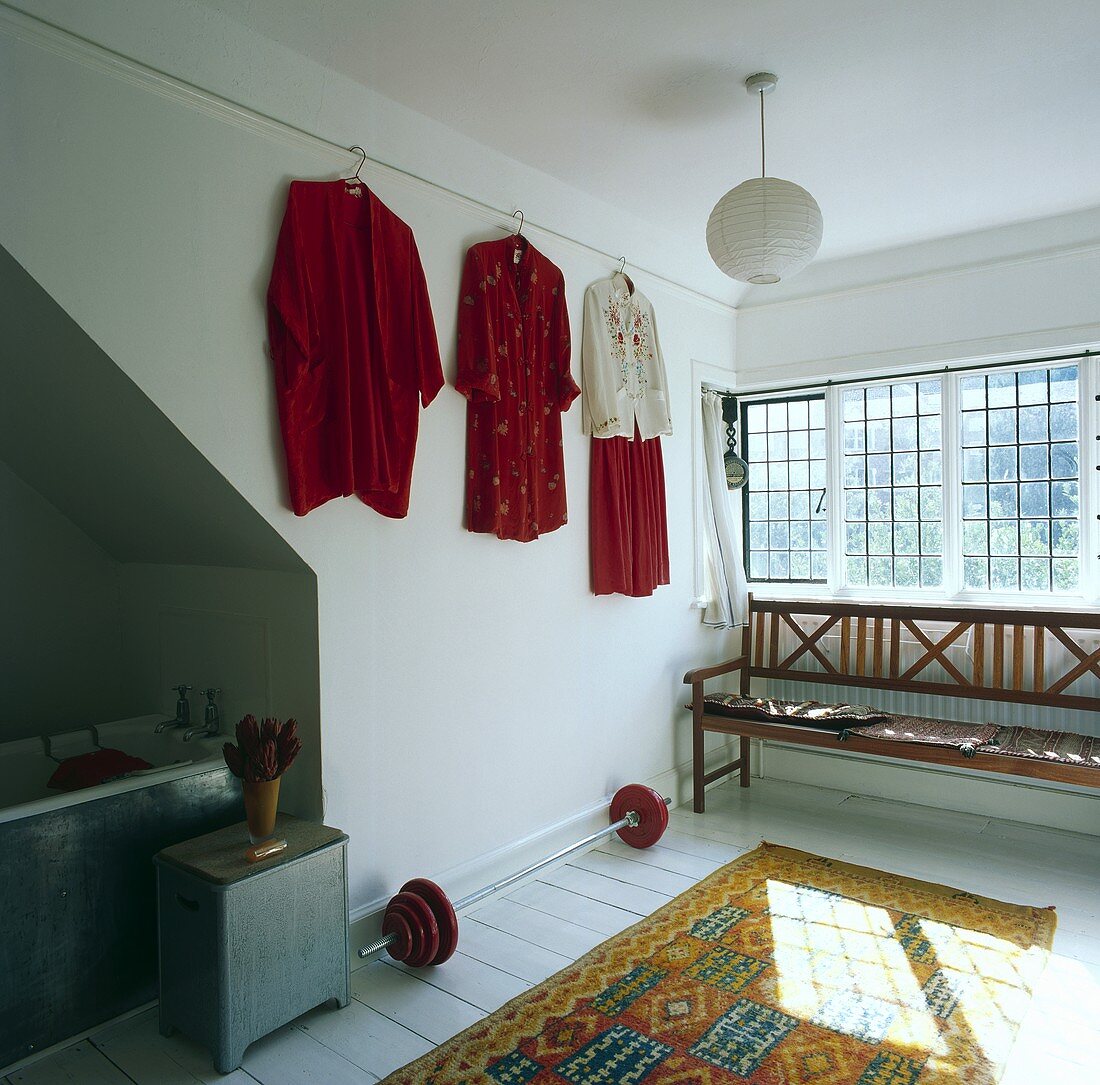 Kleider an der Wand, eine Gewichtsstange und eine Holzbank unter dem Fenster in einem Badezimmer