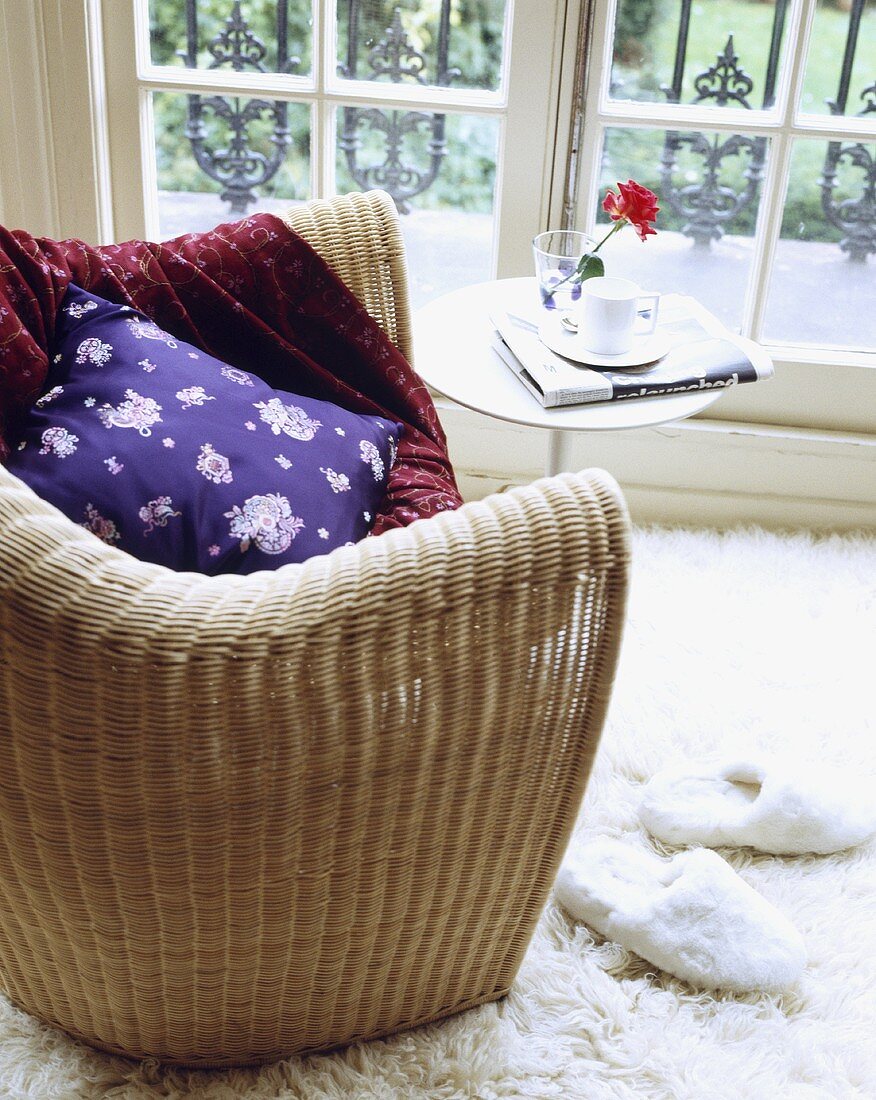 Ein Korbsessel mit lila Kissen und ein Beistelltisch vor dem Fenster