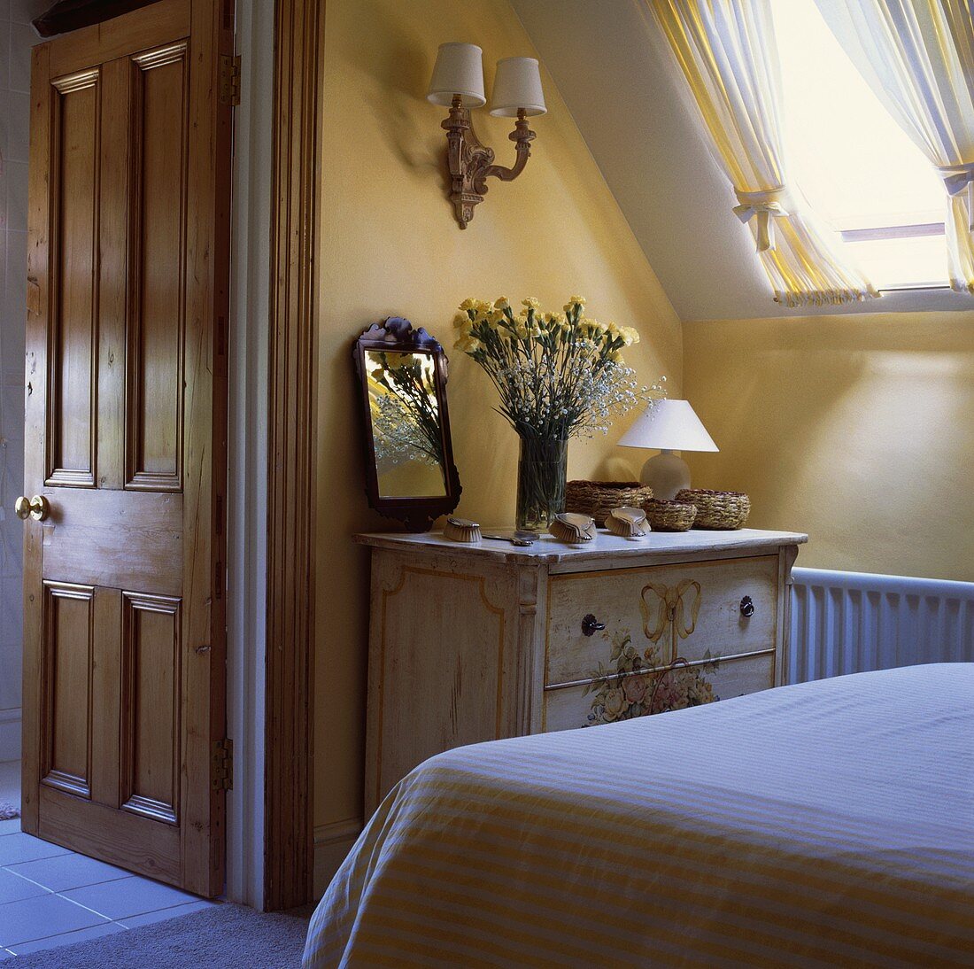 Weissliche antike Holzkommode unter Dachflächenfenster in gelb-weißem Schlafzimmer mit offener Tür zum Badezimmer ensuite