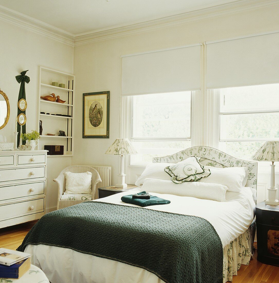 Gemütliches, weisses Landhaus-Schlafzimmer mit grünen Farbtupfern auf dem Doppelbett vor Schiebefenstern mit Stoffjalousien