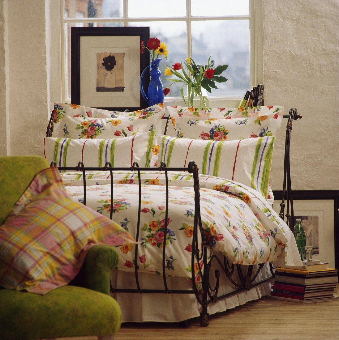 Gestreifte und geblümte Kissen mit floralem Muster auf schmiedeeisernem, antiken Bett vor Fenster mit Bild und Blumen auf Fensterbank