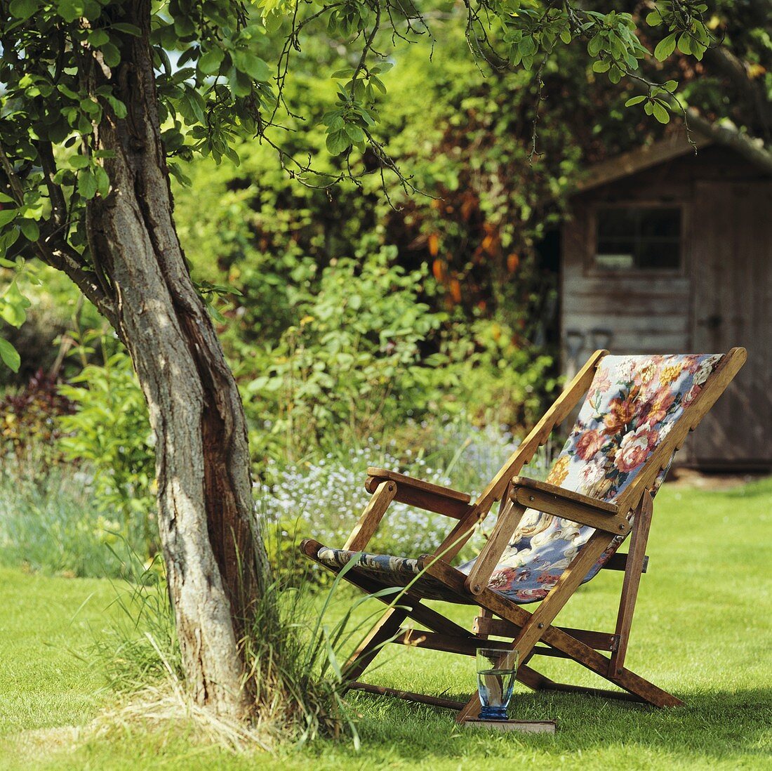 Holzliegestuhl mit blumig gemustertem Stoffbezug unter Baum in einem Sommergarten