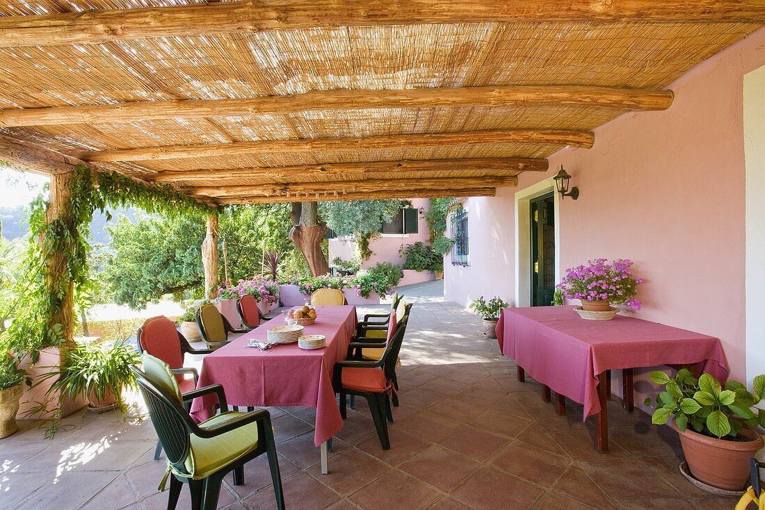 Stühle mit bunten Kissen und Tische mit rosa Decken auf der Veranda einer spanischen Villa mit Bambus-Markise