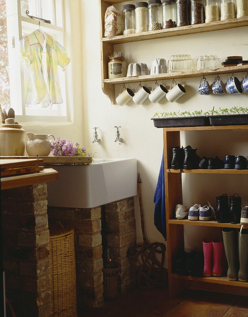 Vorratsdosen auf Wandregalen über weißem Waschbecken im Hauswirtschaftsraum mit Stiefel und Schuhen auf freistehendem hölzernen Regal gelagert