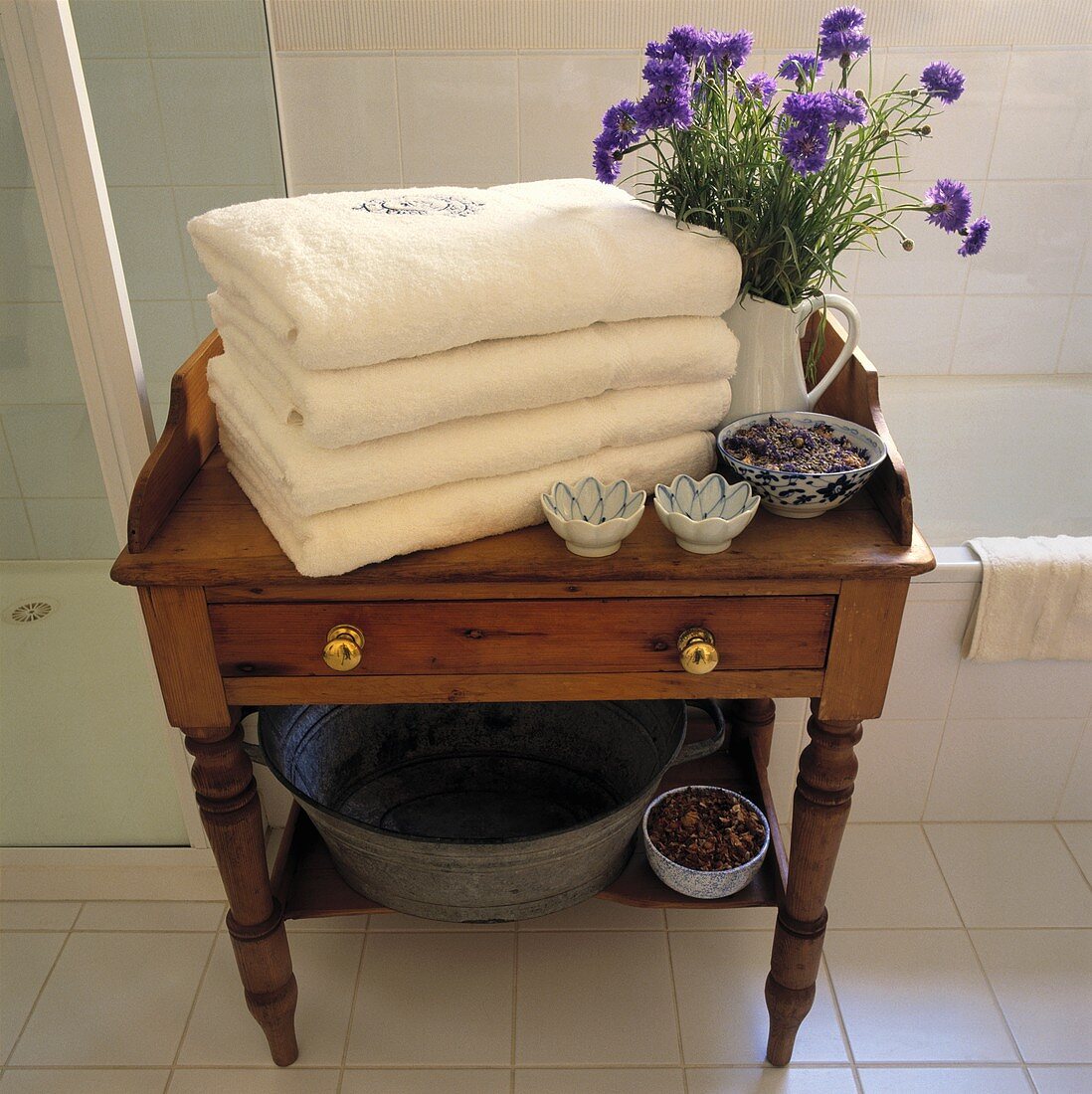 Ein Stapel von weißen Handtüchern und blauen Kornblumen in einem Krug stehen auf einen kleinen antiken Tisch aus Kiefernholz