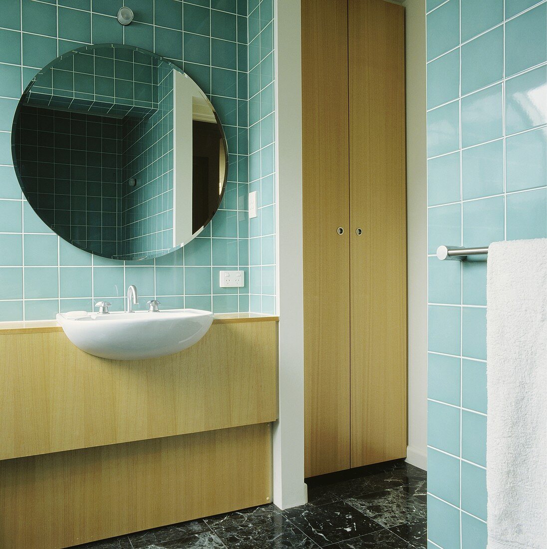 In einem türkis gefliesten Bad hängt ein kreisförmiger Spiegel über einem Waschbecken, das in einer holzgetäfelten Einheit eingelassen ist