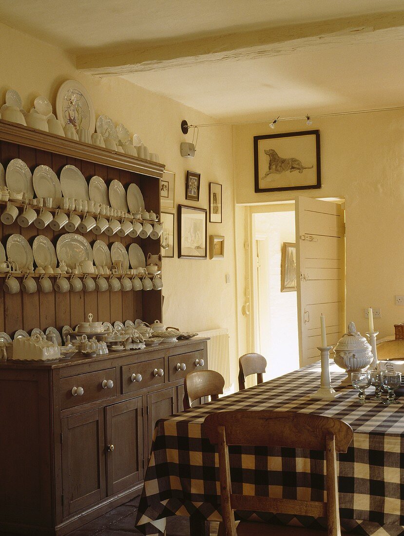 Weißes Porzellangeschirr ist in einer antiken Holzkommode aufgestellt worden, die an einer Wand der weißen Landhausküche mit einem Tisch mit schwarz-weiß karierter Tischdecke