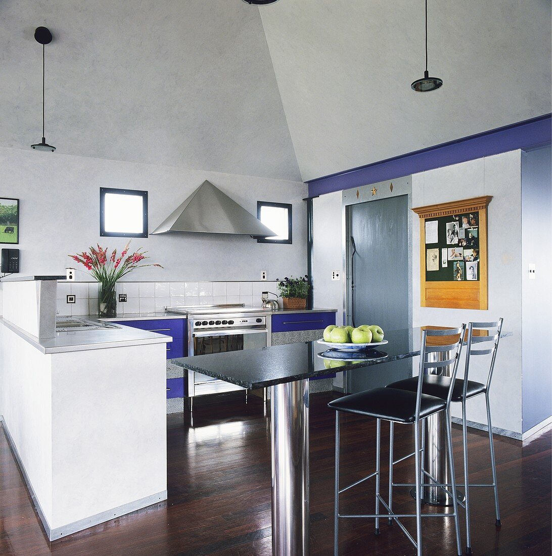 Ein Bar-Tisch, der aus einer Marmorplatte und zwei Stahlzylinder als Tischbeinen besteht, und zwei Barhocker stehen in einer modernen hellen Küche mit einem dunklen Parkettboden