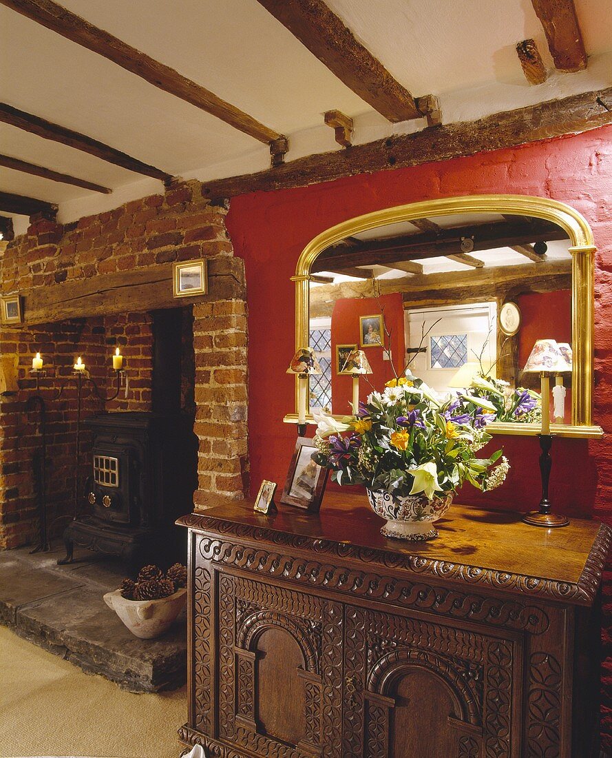 Ein vergoldeter Spiegel hängt an einer roten Wand über dem geschnitzten Schrank, der neben einer Backsteinfeuerstelle steht