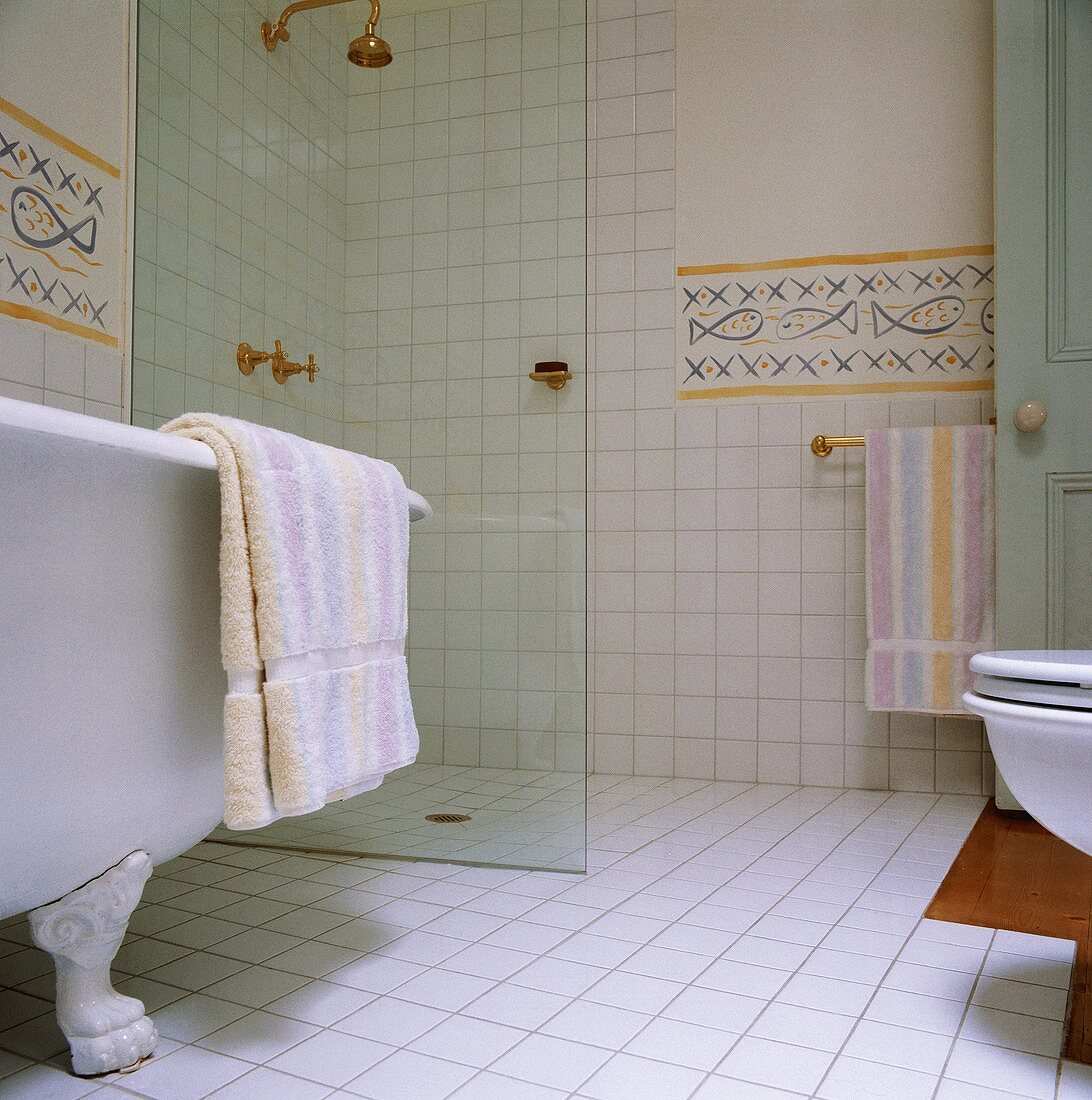 Eine freistehende Badewanne mit Klauen-Füßen steht vor der modernen Nasszelle mit einer Glaswand des weißgefliesten Badezimmers