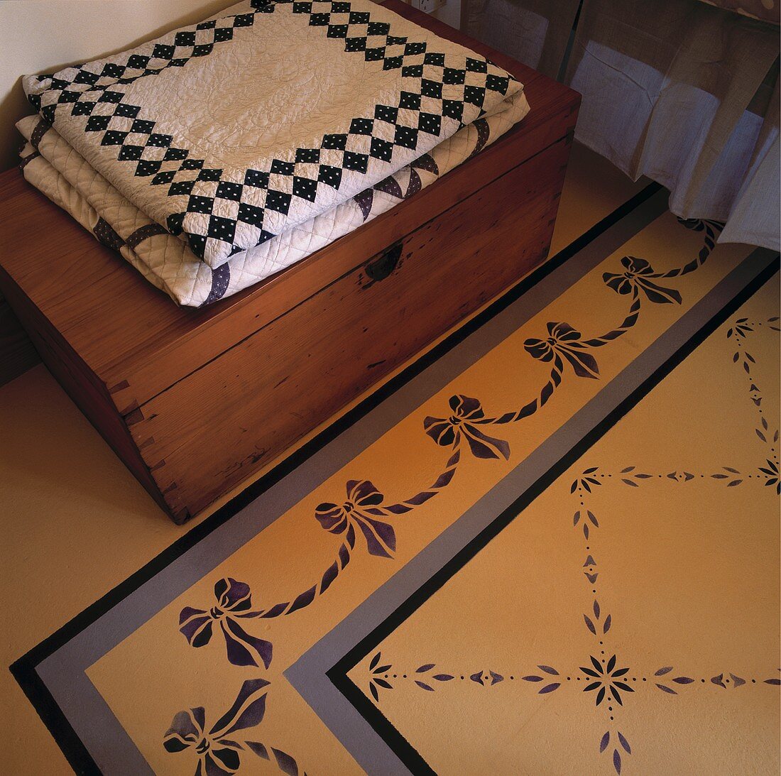 Ein Detail des illusionistischen Teppichbodens und einer Holztruhe, auf der Tagesdecken aufgestapelt worden sind