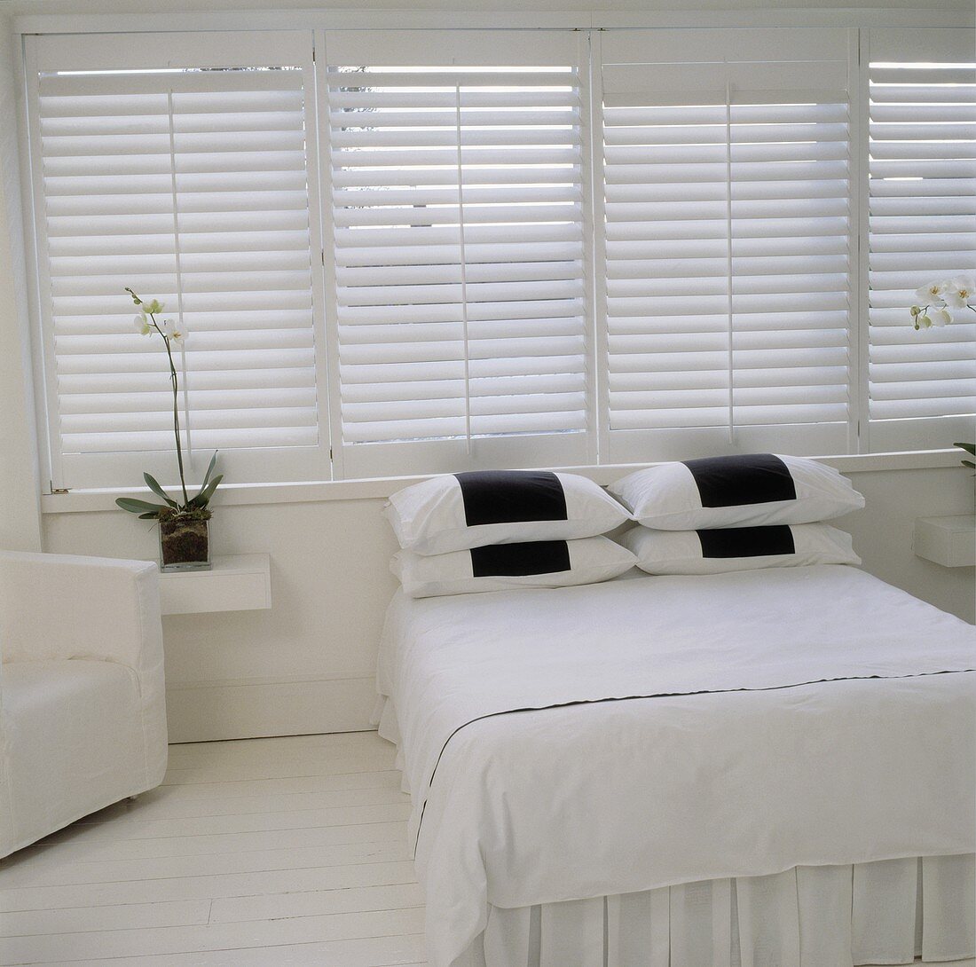 Weisses Schlafzimmer mit geschlossenen Fensterläden und Schwarzkontrast auf den kleinen Kopfkissen