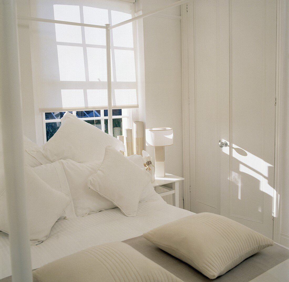 Weisses Landhaus-Schlafzimmer mit beiger Dekoration und Sonnenflecken auf Schrank und Stoffjalousie