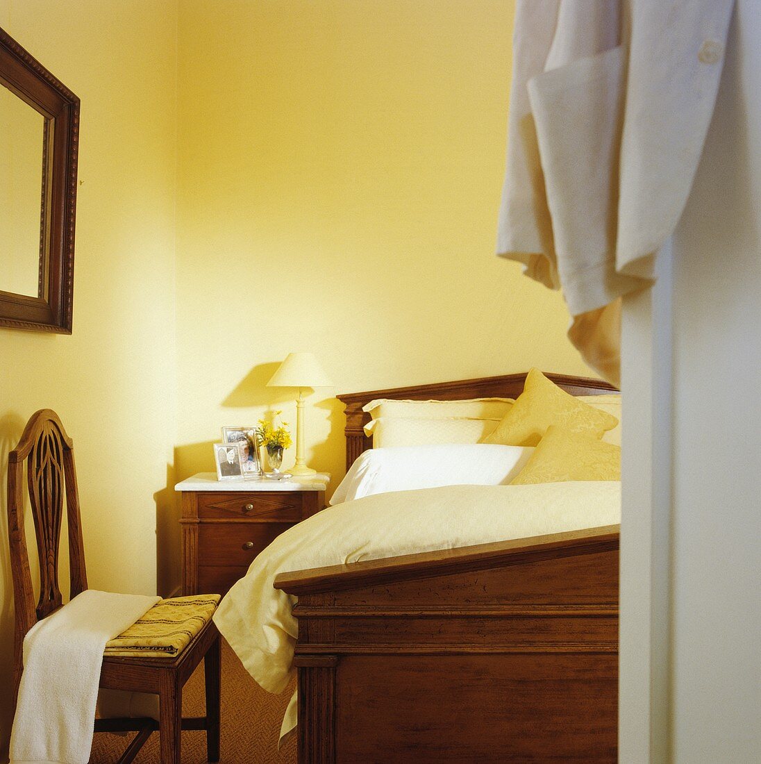 Blick durch offene Tür in pastellgelbes Schlafzimmer mit antikem Mahagoni-Bett, passendem Nachtschränkchen und Holzstuhl im Jugendstil