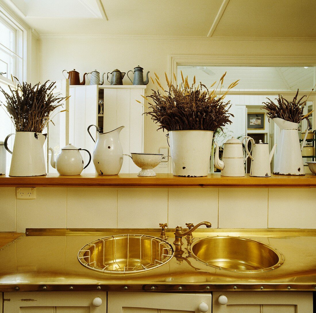 Alte, weiße Emaillekrüge auf Schrank und Küchentheke über genieteter Messingarbeitsfläche mit zwei kreisrunden Spülbecken und antiker Armatur