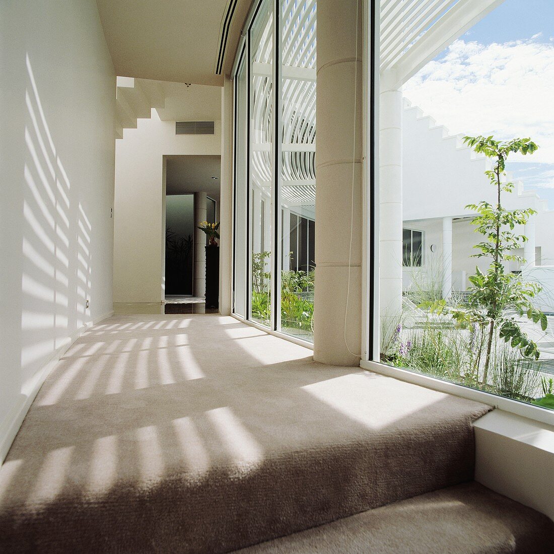 Leicht gebogene Gangflucht in modernem, weißem Haus mit hellgrauem Teppichboden und einseitiger Glasfront mit Sonnenschutz-Pergola zu Innenhof