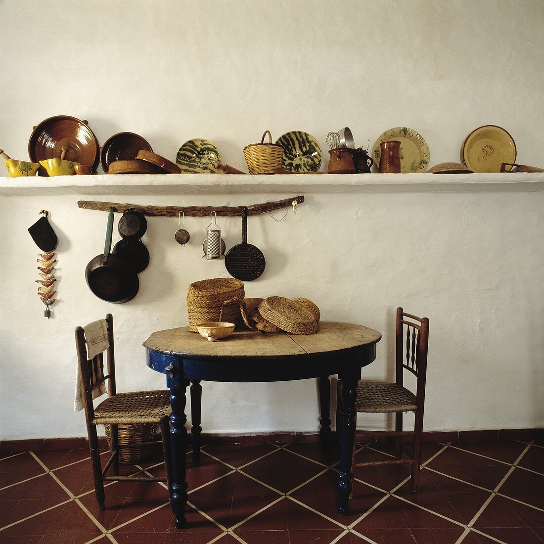 Körbe, Keramikteller und Pfannen auf Wandbord hinter rundem Holztisch mit antiken Stühlen in rustikaler Küche
