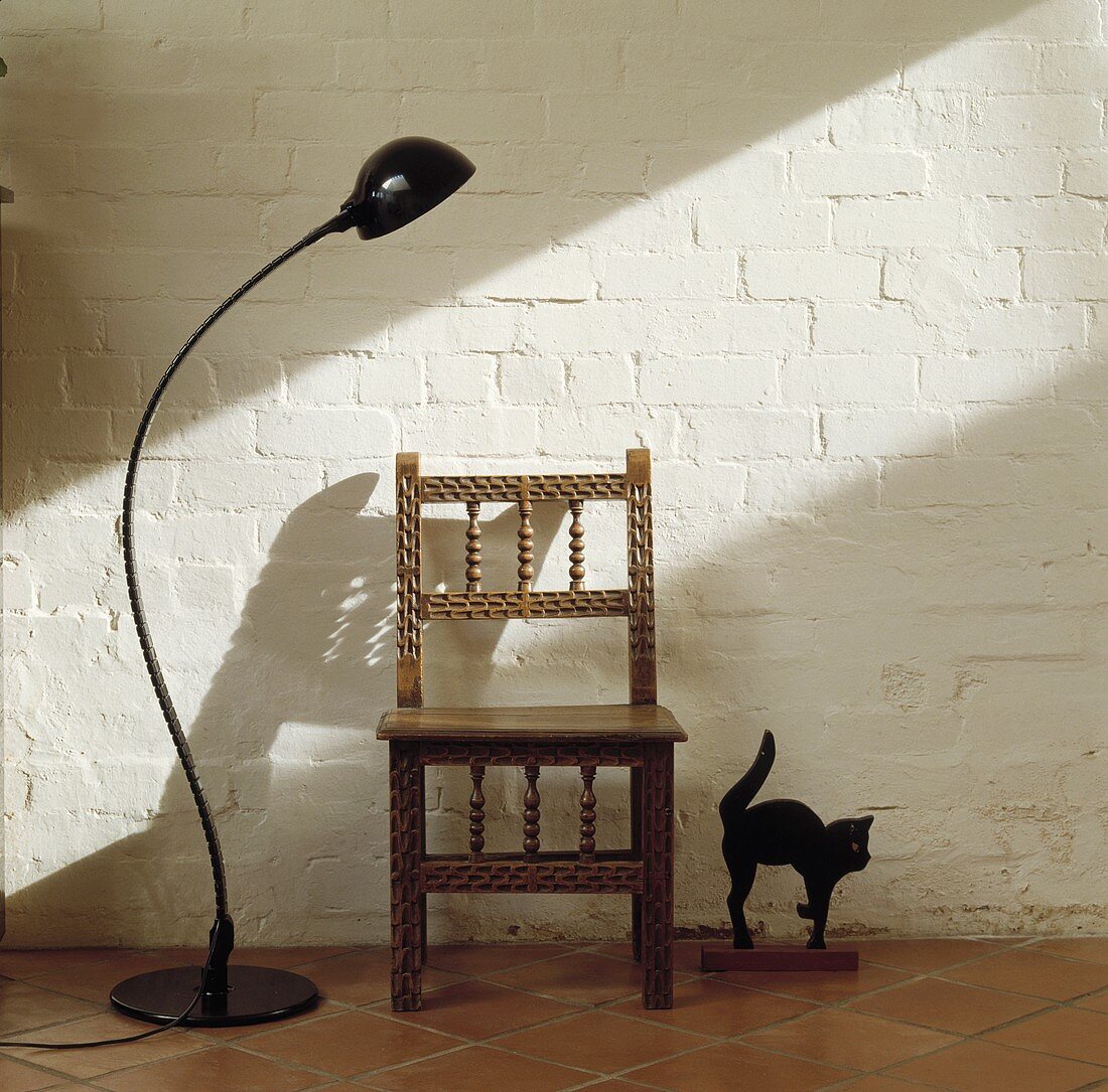 Schwarze Stehlampe und schwarze Katzenskulptur mit holzgeschnitztem Stuhl vor weiss geschlemmter Ziegelmauer