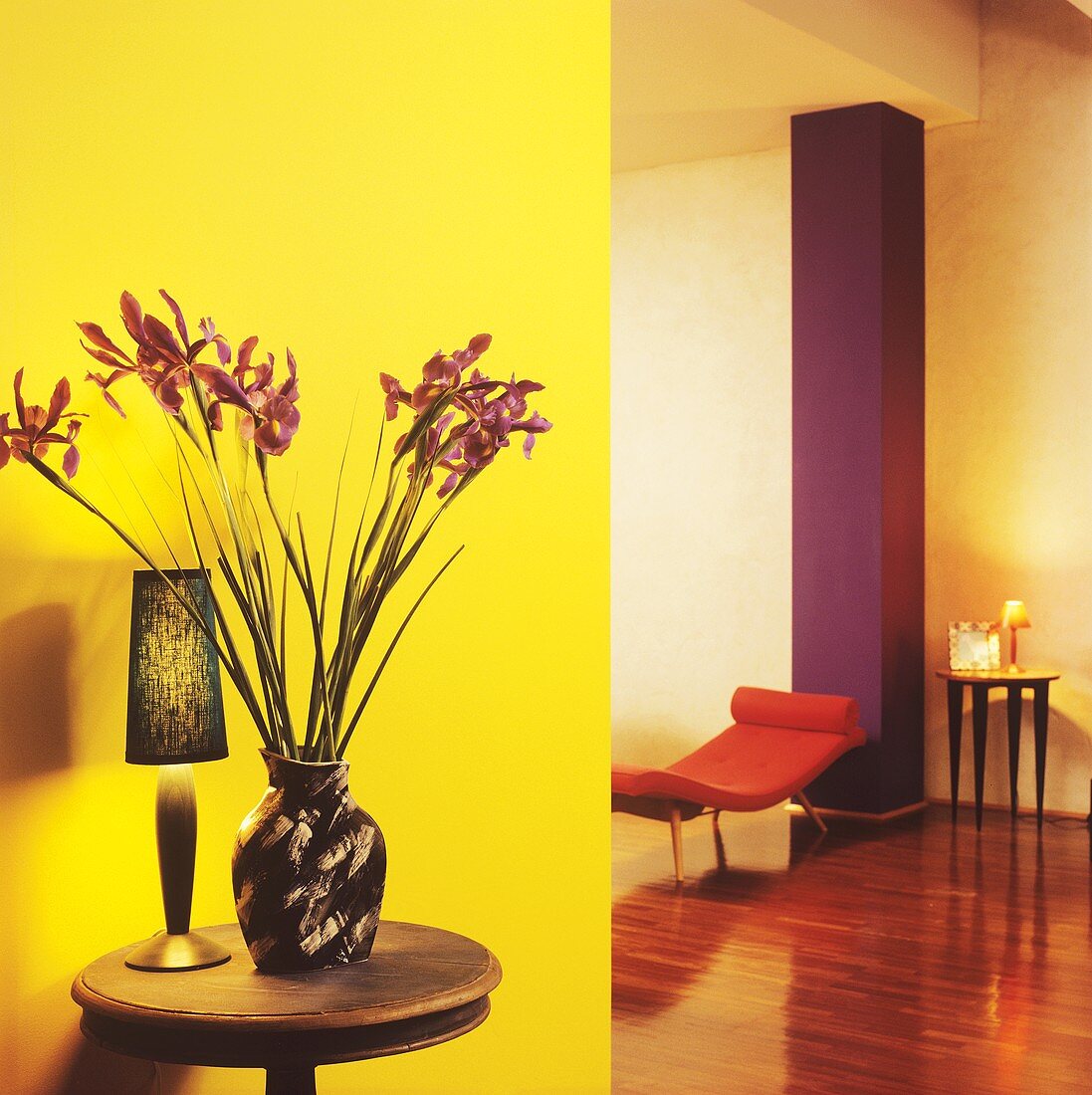 Vase mit Iris auf kleinem Holztisch vor gelber Wand in Kontrast mit lilafarbener Mauersäule und orangefarbener Liege in modernem Wohnzimmer