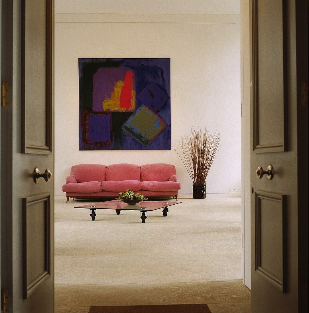 Ein Couchtisch aus Glas und ein pinkfarbenes Sofa unter einem abstrakten Bild