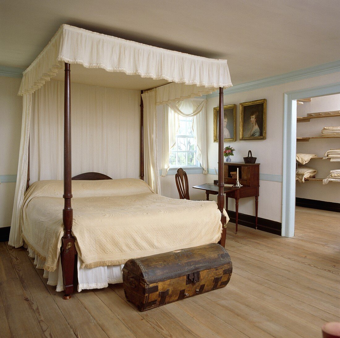 Schlafzimmer im kolonialen Stil mit cremefarbener Bettdecke auf dem Himmelbett, kleiner antiken Holztruhe und Holzboden