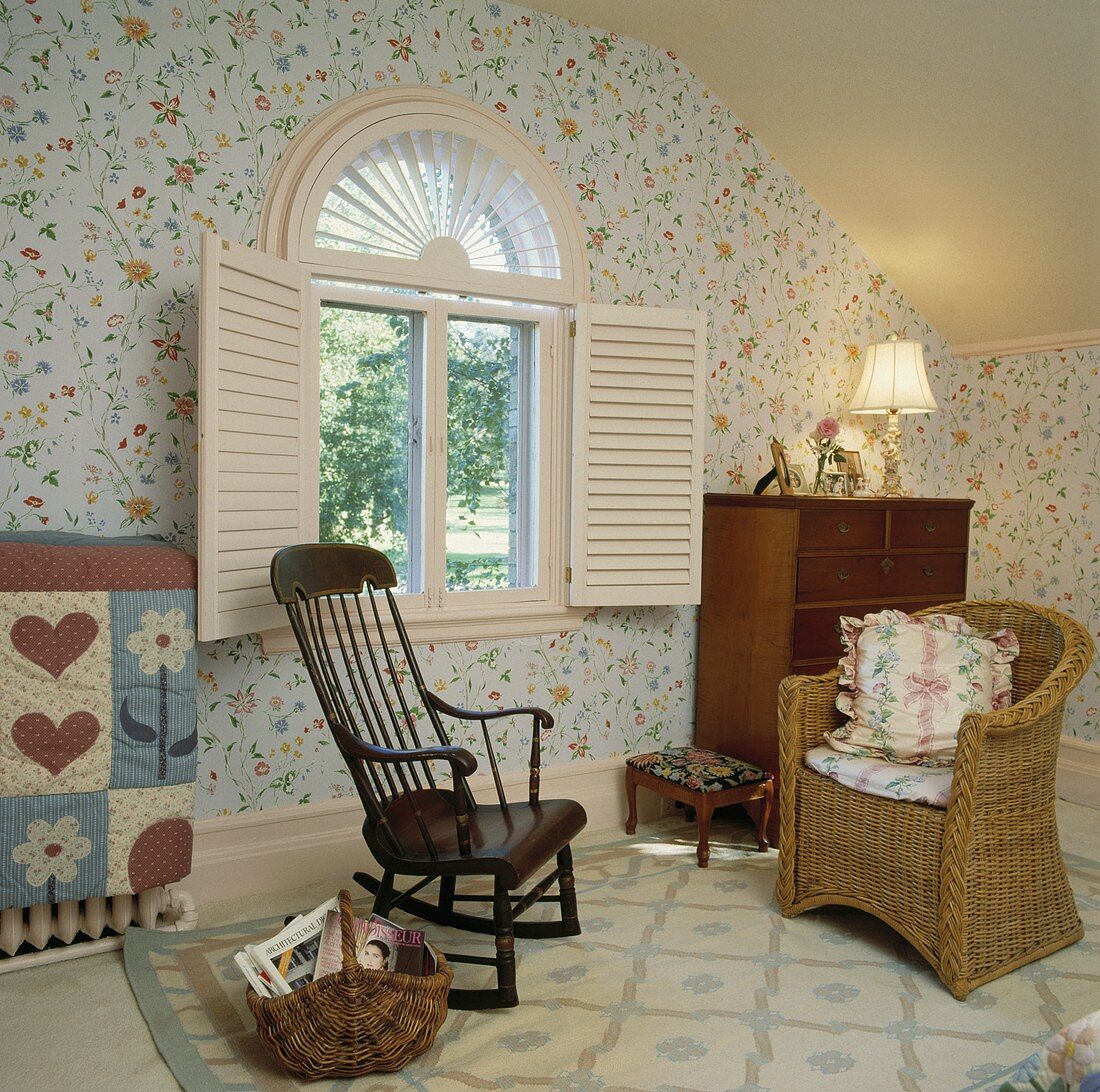 Schaukelstuhl aus Eichenholz und Korbsessel im Schlafzimmer mit floralen Tapeten und weissen Fensterläden vor einem Bogenfenster