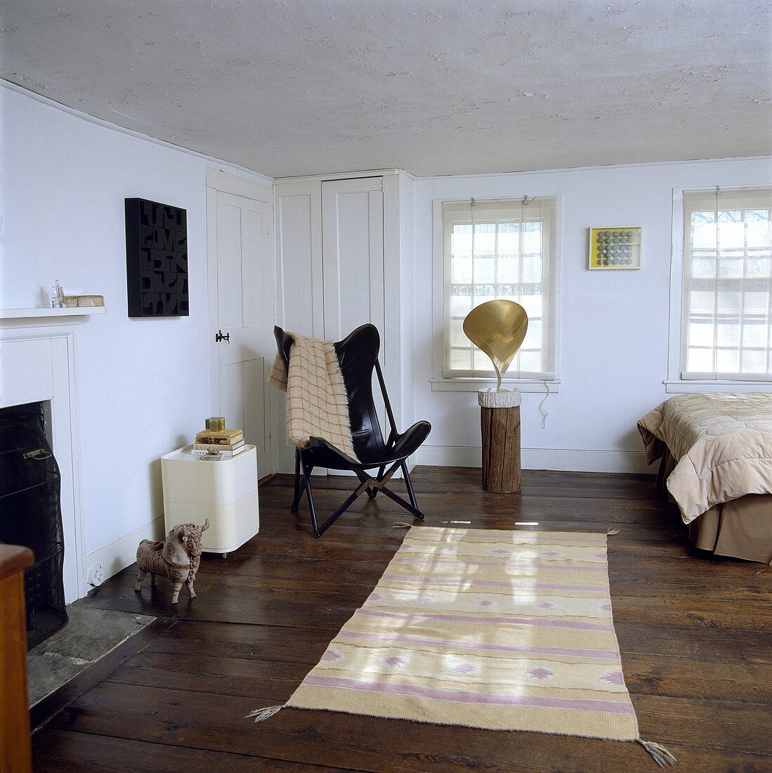 Weisses Schlafzimmer mit schwarzem Butterfly Sessel, gemustertem Ikat-Teppich, alten Holzdielen und moderner Skulptur