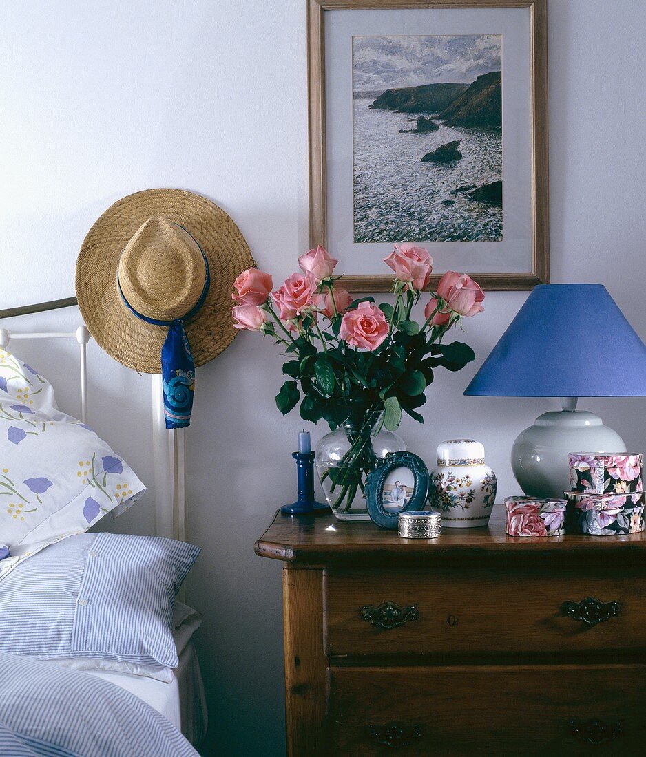 Rosen in Vase auf antik rustikalem Nachttisch und Strohhut auf Messinggestell am Kopfende vom Bett