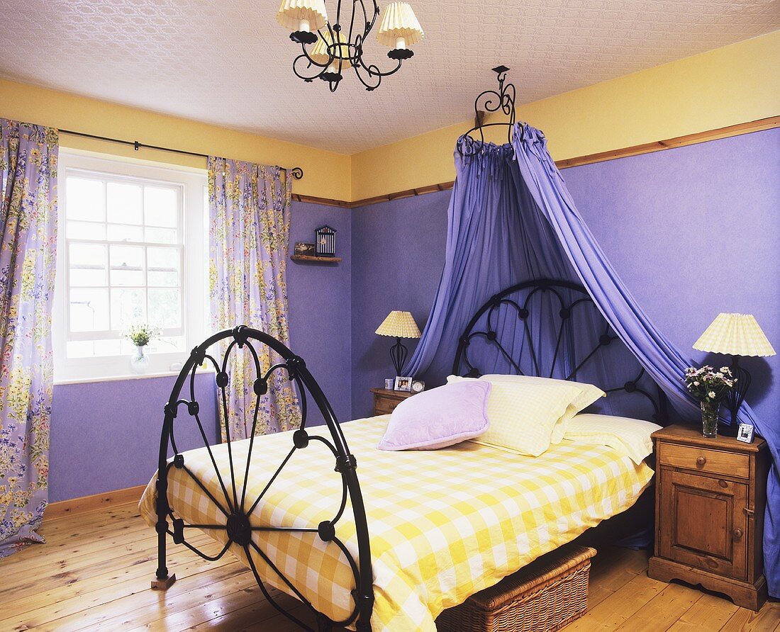 Schwarzes schmiedeeisernes Bett mit gelb weiß karierter Bettdecke und lila Baldachin vor lila Schlafzimmerwand