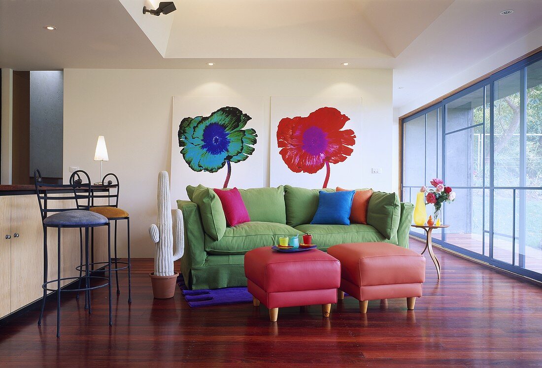 Pinkfarbene Hocker und grünes Sofa vor Blumenbilder in einem modernen Wohnzimmer mit Parkettboden