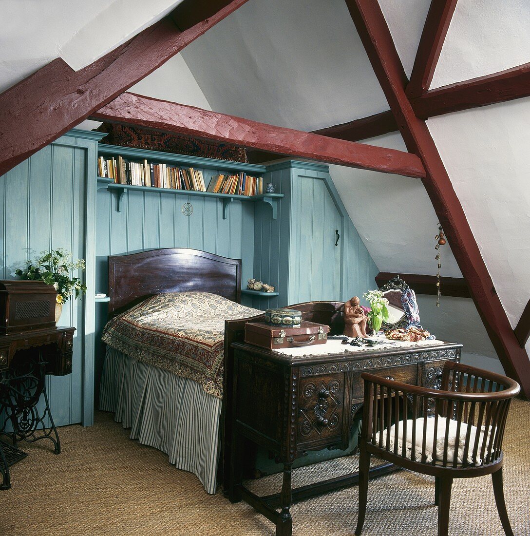 Schlafzimmer mit antikem Bett und Schreibtisch am Bettende vor blau getönter Wand unter Dach mit Dachstuhl