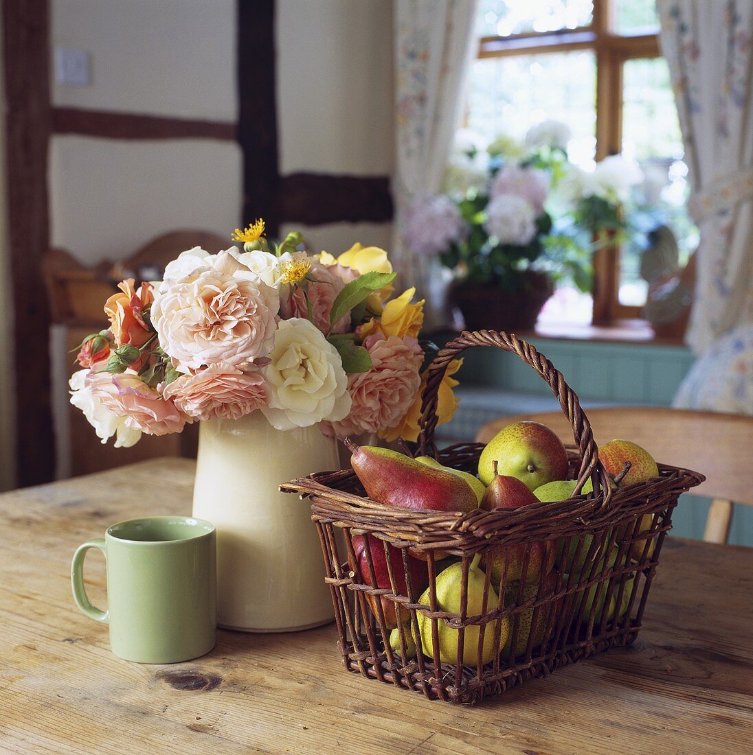 Korb mit Birnen und Rosenstrauss im Krug neben grüner Tasse auf Holztisch