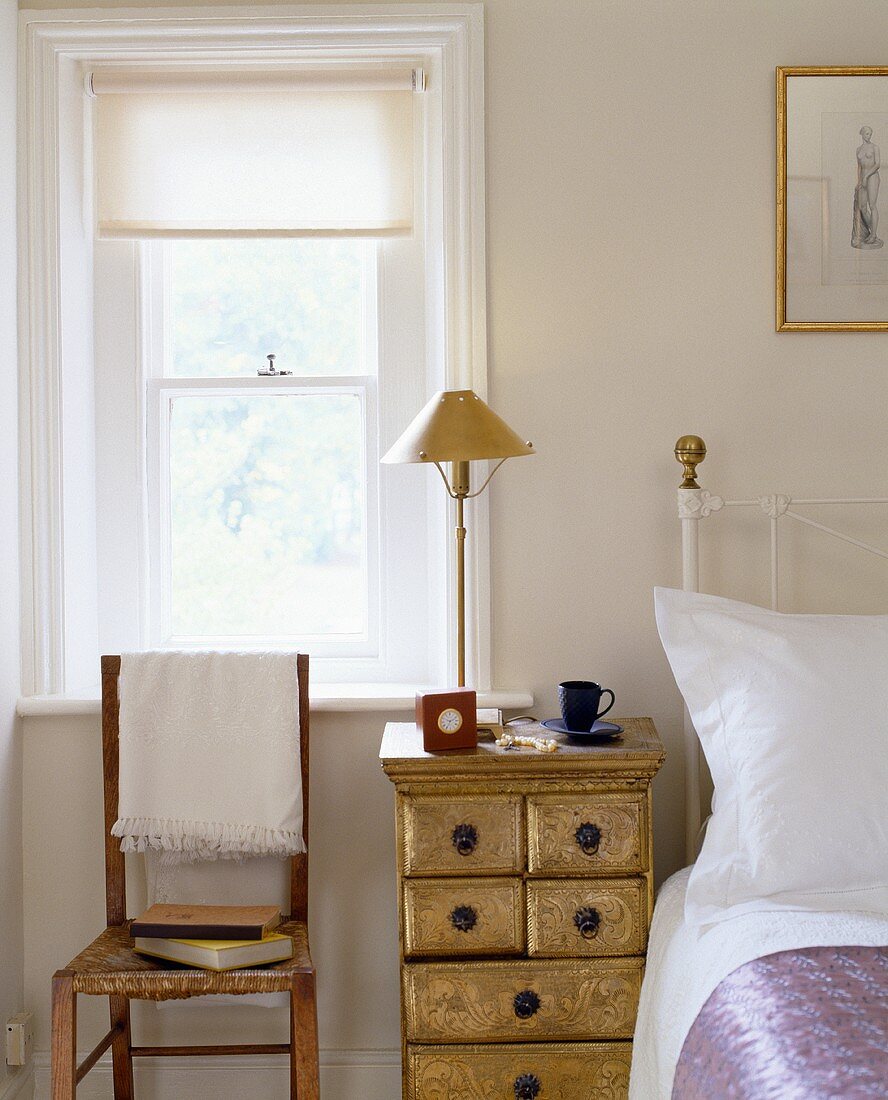Holzstuhl mit weisser Decke vor Fenster und Nachttisch mit Schubladen