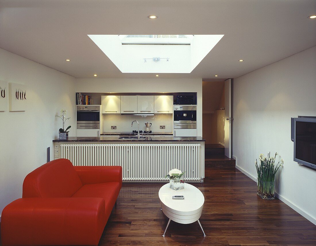 An open-plan living room with a red sofa, a white coffee table, parquet flooring and and open-plan kitchen in the background