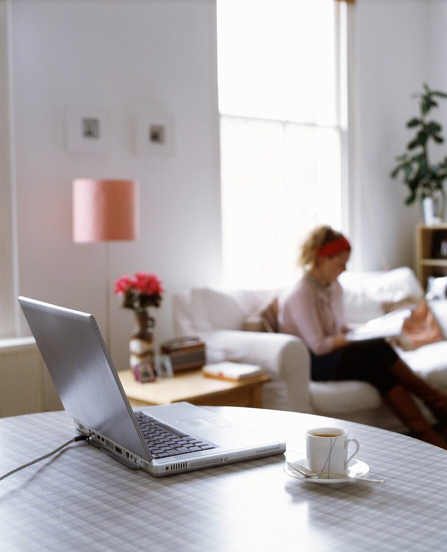 Laptop und Kaffeetasse auf Tisch, im Hintergrund Frau lesend auf dem Sofa