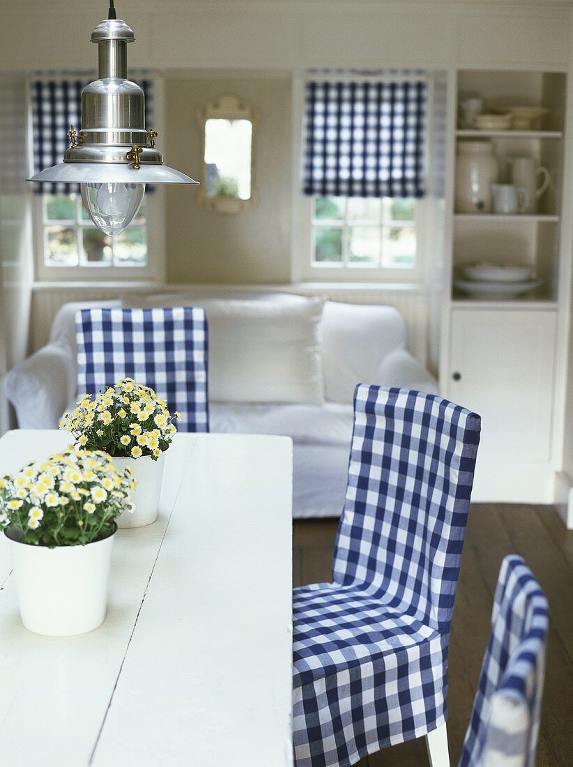 Wohnraum mit Esstisch, blau-weiss karierten Stühlen, maritimer Hängeleuchte, weißem Sofa & blau-weiss karierten Raffrollos im Hintergrund