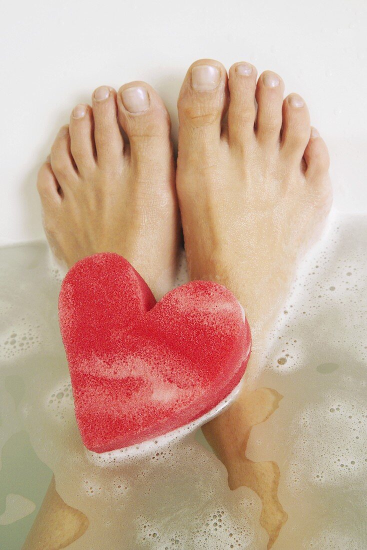 Feet and a heart-shaped sponge in a bath