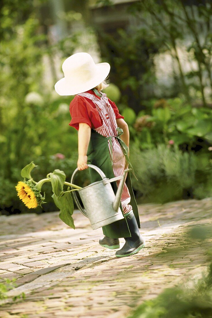 A little girl dressed as a gardner holding a watering can and a sunflower
