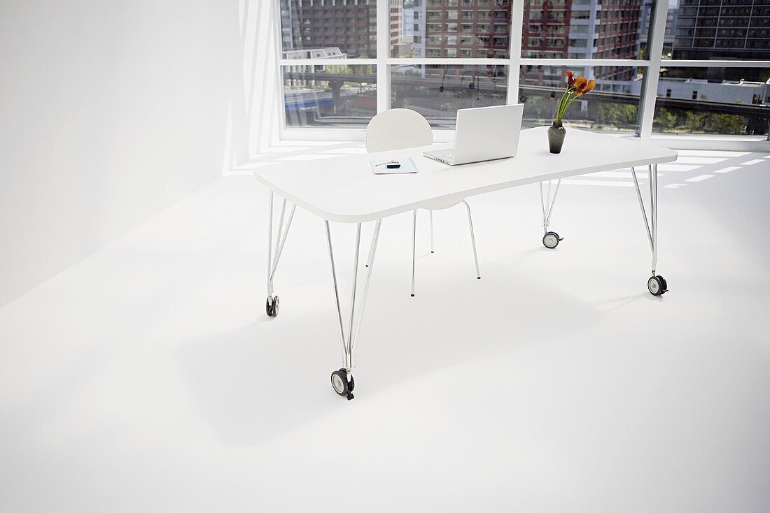 Schlichter, weißer Büroraum mit rollbarem, filigranem Schreibtisch vor großer Fensterfront
