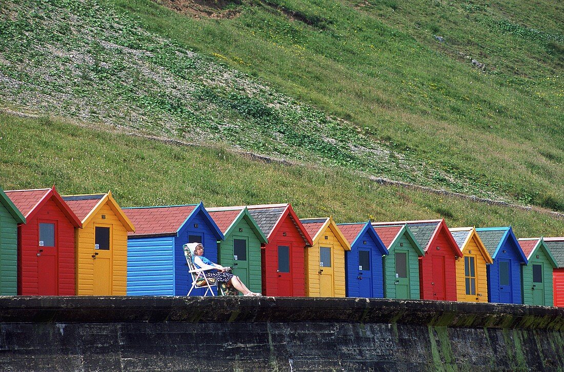 A woman sitting in a camping chair in front of colouful beach huts